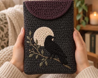 May include: A dark gray crocheted sleeve featuring a flap closure in a deep plum shade. The front displays an embroidered design of a black crow perched on a branch against a large, circular cream-colored moon, crafted with detailed stitching.