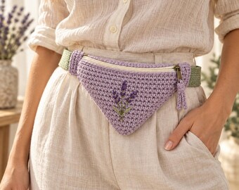 May include: A triangular crochet fanny pack in light purple featuring an embroidered lavender sprig on the front. The bag has a cream-colored zipper closure with a matching purple tassel pull and is attached to a textured olive green strap.