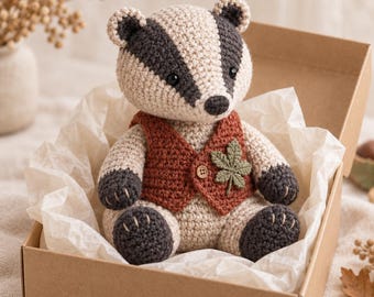 May include: A crocheted badger stuffed animal featuring a cream and dark gray striped face, dark gray paws, and a rust-colored vest. The vest is adorned with a single wooden button and a small green crocheted leaf appliqué on the left side.