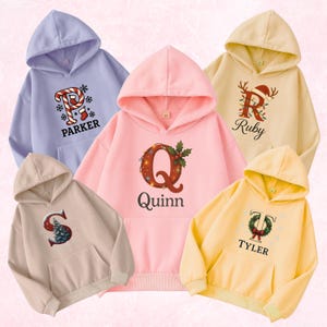 May include: Five hooded sweatshirts in pastel colors: lavender, pink, beige, yellow, and cream. Each sweatshirt features a unique Christmas-themed initial and name design. The designs include letters with festive decorations and names like "Parker" and "Quinn".