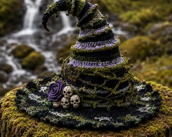May include: A crocheted witch hat featuring a spiraling design in black, olive green, and lavender yarn. The hat is adorned with a purple crocheted rose and three small white skull figures at the base, with white spiderweb embroidery on the brim.