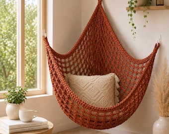 May include: A rust-colored macrame hanging chair suspended from three wall hooks, featuring a grid-like woven pattern. A square, cream-colored textured throw pillow with a diamond pattern and fringe edges is placed inside the seat of the chair.