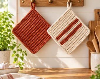 May include: Two square crochet potholders hanging on a wooden rack. One potholder is solid burnt orange with a textured waffle stitch, while the other is cream-colored with two vertical burnt orange stripes and a matching hanging loop.
