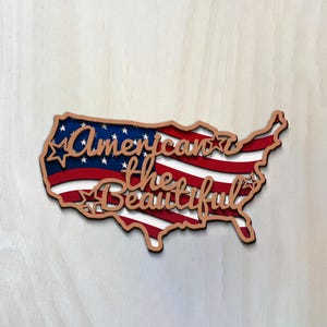 May include: Wooden cutout of the United States with the American flag design. The words "American the Beautiful" are cut out in a cursive font. The cutout is brown with red, white, and blue stripes and stars.