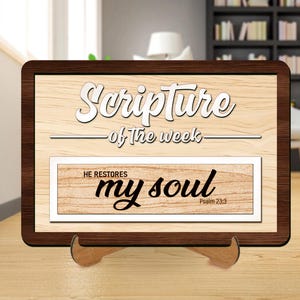 Op de afbeelding: Houten bord met de woorden "Scripture of the week" in witte letters. Daaronder staat in een kleinere rechthoek "He restores my soul" met "Psalm 23:3" in de hoek. Het bord wordt op een houten standaard gepresenteerd.