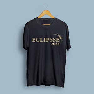Peut inclure: T-shirt noir avec l'inscription "ECLIPSE 2024" en or. Le design comprend un soleil stylis&eacute; ou un motif d'&eacute;clat d'&eacute;toile. Le t-shirt est suspendu &agrave; un cintre en bois sur fond bleu clair.