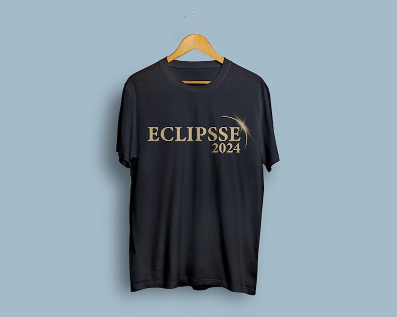 Peut inclure: T-shirt noir avec l'inscription "ECLIPSE 2024" en or. Le design comprend un soleil stylis&eacute; ou un motif d'&eacute;clat d'&eacute;toile. Le t-shirt est suspendu &agrave; un cintre en bois sur fond bleu clair.