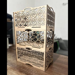 Könnte beinhalten: Ein dreistufiges Holzregal mit aufwendigen Laser-Cut-Designs. Jede Ebene weist ein anderes Muster auf, darunter florale, geometrische und Gittermuster. Das Regal hat ovale Griffe und ist aus hellem Holz gefertigt, geeignet zum Organisieren von Gegenständen.