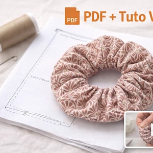 以下が含まれることがあります： ピンクと茶色の花柄のシュシュが、糸巻き、安全ピン、パターンと一緒に白い表面に置かれています。画像には「PDF + Tuto Vidéo」というテキストも含まれています。