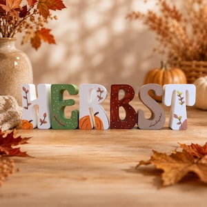 Könnte beinhalten: Ein Set aus sechs freistehenden Holzbuchstaben, die das Wort HERBST bilden. Jeder Buchstabe ist mit einzigartigen saisonalen Motiven verziert, darunter gemalte orangefarbene Kürbisse, braune und grüne Blätter sowie weiße Punkte auf weißem, grünem und braunem Untergrund.