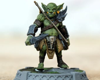 Pode incluir: Uma figura de goblin verde com uma lança e uma espada, vestindo armadura e um tapa-sexo. A figura em miniatura está em uma base cinza decorativa. O goblin tem orelhas pontudas e uma expressão ameaçadora.