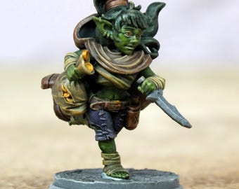 Pode incluir: Uma figura em miniatura de um personagem goblin de pele verde, segurando uma faca e um saco. O goblin usa uma capa marrom, um cinto e calções roxos. A figura é montada em uma base cinza texturizada.