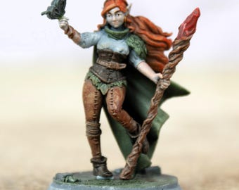 Pode incluir: Figura de fantasia em miniatura de uma elfa com cabelo ruivo, vestindo uma capa verde, um espartilho marrom e leggings. Ela segura uma bengala com ponta vermelha e uma borboleta. A figura está em uma base cinza.