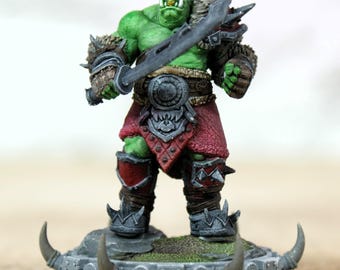 Pode incluir: Figura em miniatura detalhada de um guerreiro orc de pele verde, segurando uma grande espada. O orc está vestido com uma armadura vermelha e marrom com detalhes de pele. A figura está em uma base cinza texturizada com protuberâncias em forma de chifre, com tema de fantasia.