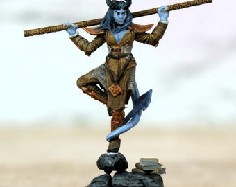 Pode incluir: Figura de fantasia em miniatura de um humanoide de pele azul com chifres, segurando um bastão. A figura usa uma roupa marrom e dourada e fica em uma base rochosa com pequenos livros. A figura tem aproximadamente 7.6 cm de altura.