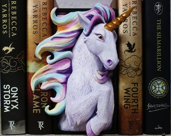 Pode incluir: Um suporte de livros de unicórnio caprichoso, com um unicórnio cor de lavanda com um chifre dourado e uma crina de cores pastel. O suporte de livros está posicionado entre livros com títulos como "Onyx Storm" e "Fourth Wing".