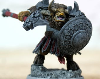 Pode incluir: Miniatura de um guerreiro minotauro, pintada em tons de castanho, cinza e vermelho. O minotauro está posando com um escudo e uma arma, em pé sobre uma base texturizada. A miniatura tem armadura detalhada e uma expressão feroz.