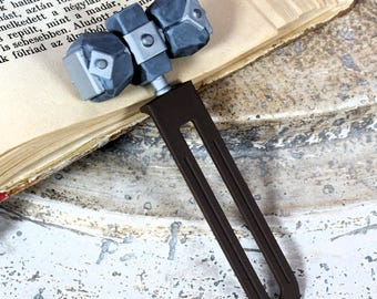 Pode incluir: Um marcador de livros único com design de martelo. A cabeça do martelo é cinza com detalhes semelhantes a pedra, conectada a uma longa alça marrom escura. O marcador de livros está nas páginas de um livro aberto.