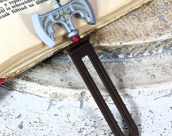 Pode incluir: Um marcador de livros decorativo em forma de machado, com uma lâmina prateada e dourada e um cabo castanho. O marcador de livros está colocado nas páginas de um livro aberto. Um acessório único para amantes de livros.