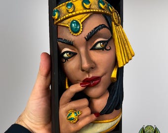 Könnte beinhalten: Gerahmte Reliefskulptur einer Frau mit dunklem Haar und goldener Kopfbedeckung. Die Frau hat roten Lippenstift, einen goldenen Ring und berührt ihre Lippen mit einem Finger. Die Skulptur ist ein Porträt einer Frau mit geheimnisvollem Ausdruck.