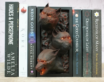 Könnte beinhalten: Ein dekoratives Bücherregal mit einer dunklen, dreidimensionalen Skulptur von knurrenden Wolfsköpfen mit leuchtend orangefarbenen Augen. Die Skulptur befindet sich zwischen mehreren Büchern mit Titeln wie "Hades & Persephone" und "Neon Gods".