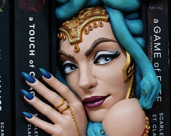 Pode incluir: Uma mulher com olhos azuis, batom roxo escuro e unhas azuis, adornada com um adorno de cabeça dourado e um adorno de cabeça em forma de cobra. A imagem está ao lado de uma pilha de livros com os títulos "a TOUCH of MALICE" e "a GAME of FATE".
