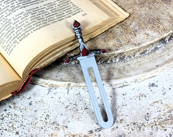 Pode incluir: Um marcador de livro prateado em forma de espada com detalhes vermelhos repousa sobre um livro aberto. O marcador de livro apresenta uma empunhadura detalhada e uma lâmina longa e fina. As páginas do livro estão envelhecidas.