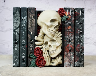 May include: A decorative bookend featuring a pair of skulls embracing, with red roses and green leaves. The bookend holds several books with titles like "Hunting Adeline" and "Blood of my Monster". The books are dark with white text.