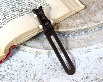 Pode incluir: Um marcador de livros castanho em forma de guitarra com pinos de afinação brancos repousa sobre um livro aberto. O marcador de livros é feito de um material castanho escuro e tem um corpo longo e fino. O livro tem páginas envelhecidas e uma lombada vermelha.