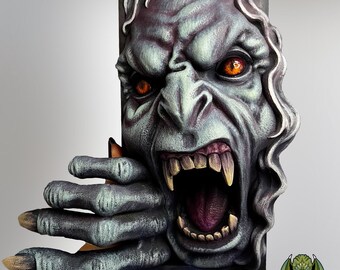 Pode incluir: Escultura pintada à mão de um rosto monstruoso com dentes afiados e olhos laranja brilhantes. A mão da criatura estende-se de uma moldura escura, criando um efeito 3D. A paleta de cores inclui tons de cinza, roxo e verde, com um toque de branco.