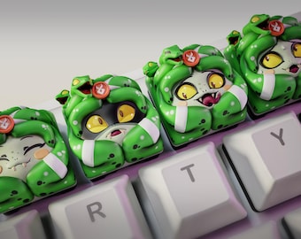 Könnte beinhalten: Vier individuelle Tastenkappen mit einem Cartoon-Medusa-Design. Die Tastenkappen haben grüne Schlangen als Haare, gelbe Augen und einen roten Knopf. Die Tastenkappen befinden sich auf einer weißen Tastatur, auf der die Buchstaben R, T und Y zu sehen sind. Das Nom Nom Figures-Logo befindet sich oben links.