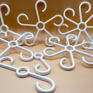 Könnte beinhalten: Eine Sammlung weißer Kunststoff-Mehrfachkleiderbügel, die jeweils einen zentralen kreisförmigen Ring mit sechs strahlenförmigen Armen aufweisen, die in gebogenen Haken enden. Diese Organizer sind zum Aufhängen mehrerer Gegenstände wie Gürtel oder Schals gedacht.