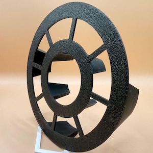 Könnte beinhalten: Ein schwarzes, kreisförmiges Objekt mit strukturierter Oberfläche und einem radartigen Design mit acht Speichen, die von einem zentralen Ring ausgehen. Das Objekt hat gebogene, flossenartige Vorsprünge auf der Rückseite und steht auf einem weißen Ständer.