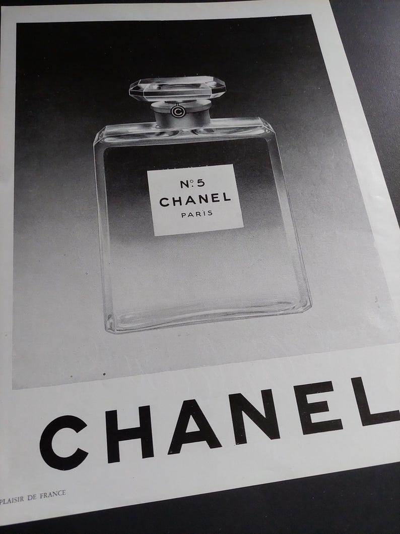 Puede incluir: Anuncio en blanco y negro que presenta un frasco de perfume. El frasco es rectangular con un cuerpo de vidrio transparente y una tapa geom&eacute;trica. La etiqueta dice "N&deg;5 CHANEL PARIS". La marca "CHANEL" se muestra debajo.