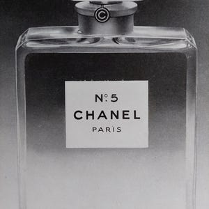 Puede incluir: Imagen en blanco y negro de un frasco de perfume. El frasco es rectangular con un tap&oacute;n facetado. La etiqueta dice "N&deg;5 CHANEL PARIS". El frasco tiene un cuello plateado y un cuerpo de vidrio transparente.