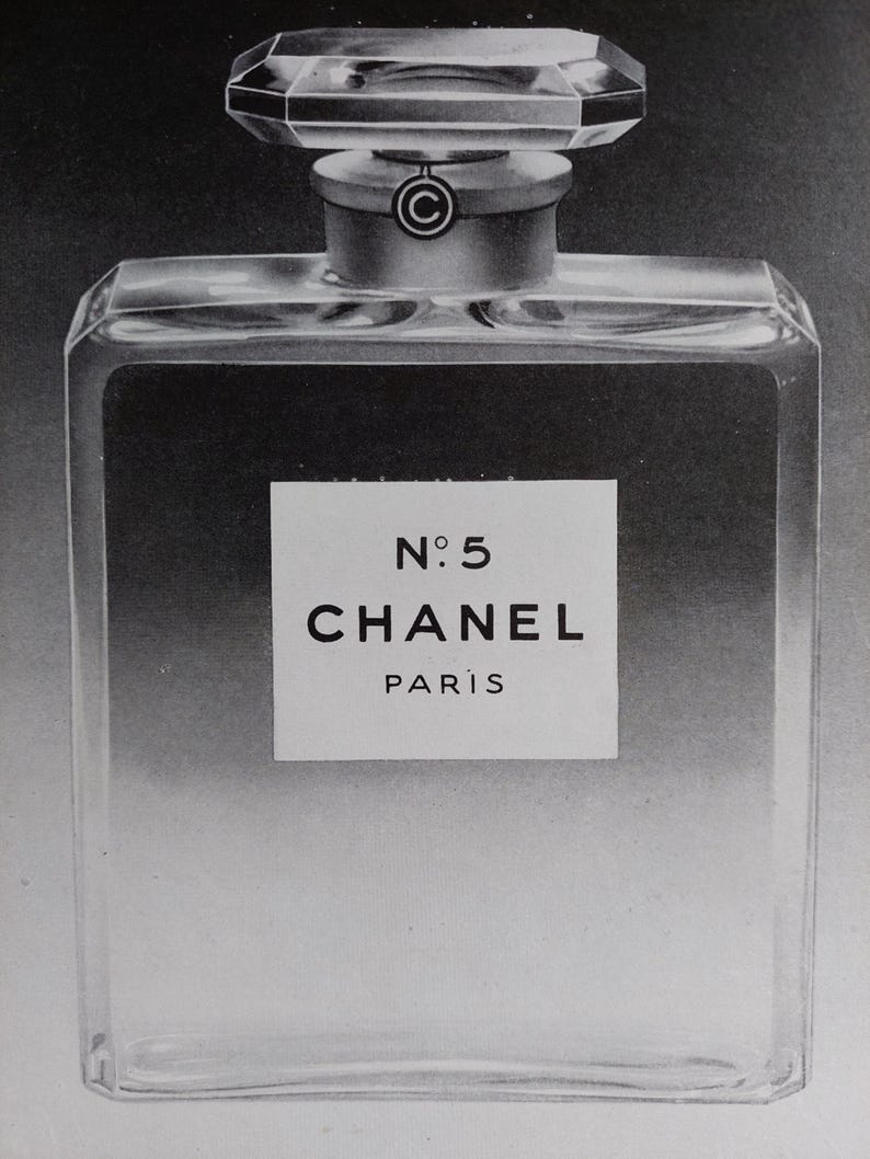 Puede incluir: Imagen en blanco y negro de un frasco de perfume. El frasco es rectangular con un tap&oacute;n facetado. La etiqueta dice "N&deg;5 CHANEL PARIS". El frasco tiene un cuello plateado y un cuerpo de vidrio transparente.