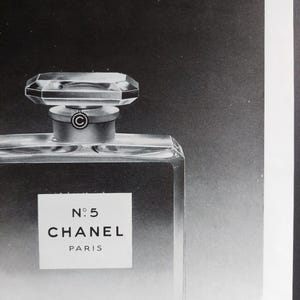 Puede incluir: Imagen en blanco y negro de un frasco de perfume. El frasco es rectangular con un cuerpo de vidrio transparente y un tap&oacute;n geom&eacute;trico facetado. La etiqueta dice "N&deg;5 CHANEL PARIS". El fondo es un degradado de negro y gris.