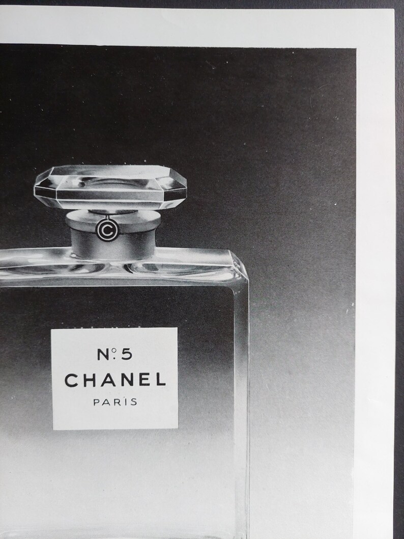 Puede incluir: Imagen en blanco y negro de un frasco de perfume. El frasco es rectangular con un cuerpo de vidrio transparente y un tap&oacute;n geom&eacute;trico facetado. La etiqueta dice "N&deg;5 CHANEL PARIS". El fondo es un degradado de negro y gris.