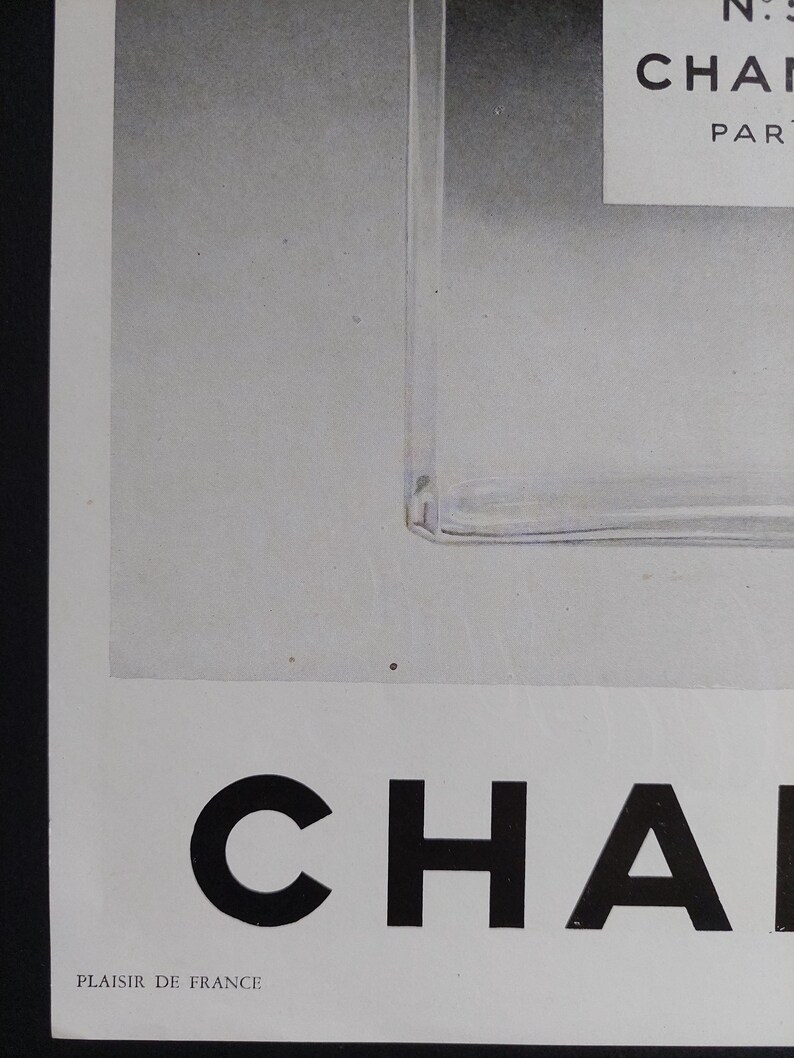 Puede incluir: Anuncio en blanco y negro con un frasco de perfume y la palabra "CHANEL" en negrita. Tambi&eacute;n se ve el texto "N:5" y "PAR". La frase "PLAISIR DE FRANCE" est&aacute; impresa en la parte inferior.