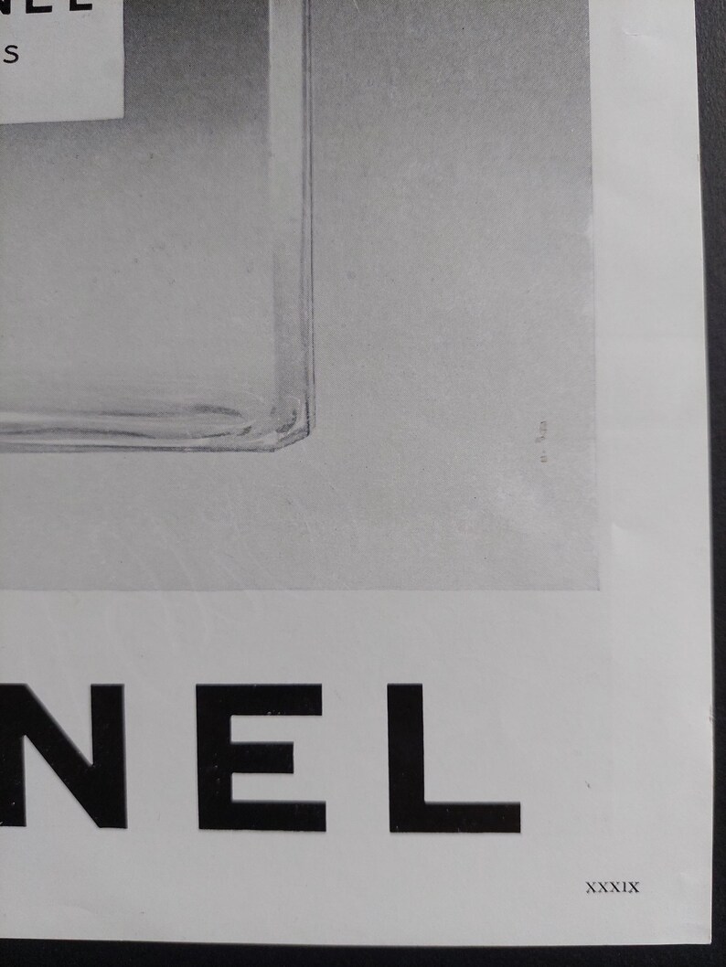 Puede incluir: Un anuncio en blanco y negro con un frasco de perfume y la palabra "NEL" en letras may&uacute;sculas negras y en negrita. La botella es parcialmente visible, con una etiqueta blanca. El n&uacute;mero romano "XXXIX" est&aacute; en la esquina inferior derecha.