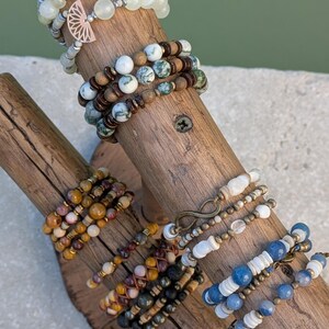 Peut inclure: Une collection de bracelets de perles multicouches présentés sur un support en bois. Les bracelets comportent diverses pierres naturelles, notamment des perles vertes, bleues, brunes et blanches, agrémentées de breloques métalliques en forme d'éventail et d'infini.