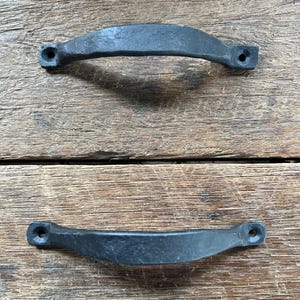 May include: Two black, curved metal handles with a textured finish. Each handle has two mounting holes on the ends. The handles are displayed on a weathered, wooden surface.