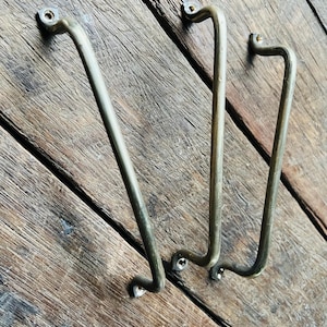 May include: Three antique brass cabinet pulls with a curved handle design. The pulls are mounted on a weathered wooden surface, showcasing their aged patina and vintage aesthetic. Each pull has a rectangular mounting plate.