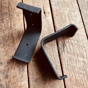 May include: Two black metal shelf brackets with a curved design. The brackets are made of a thick, sturdy metal and have a matte finish. They are designed to be mounted on a wall and support a shelf. The brackets are shown on a wooden surface.