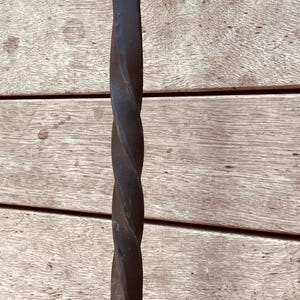 Könnte beinhalten: Ein schwarzer, gedrehter Türgriff aus Eisen. Der Griff hat eine rechteckige Basis und eine gebogene Oberseite mit einem einzelnen Loch zur Befestigung. Das gedrehte Design fügt dem Griff ein dekoratives Element hinzu.