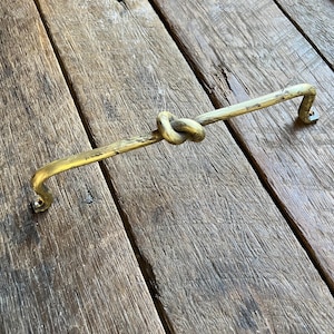 May include: A brass cabinet pull with a knot design. The handle is a warm gold colour and has a slightly textured surface. The pull is mounted on a weathered wooden surface.