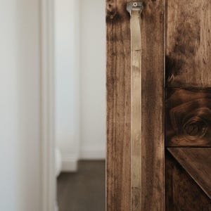 May include: Close-up of a wooden door with a long, rectangular metal handle. The door is made of dark brown wood with visible grain and knots. The handle is a light, metallic colour and is attached with screws.