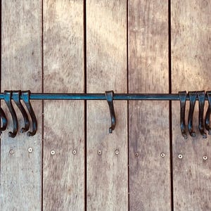 May include: A wrought iron wall-mounted coat rack with multiple curved hooks. The dark metal rack is mounted on a wooden surface with vertical planks. The hooks are evenly spaced along the horizontal bar.