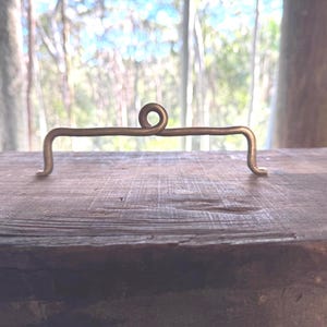 May include: A brass-coloured metal hook, handcrafted with a horizontal bar and a central looped design. The ends are curved. It rests on a dark, weathered wooden surface. The background is a blurred view of nature.
