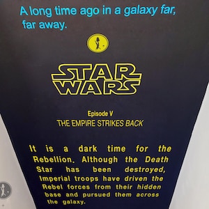 May include: A black and yellow sign with the text "Star Wars Episode V: The Empire Strikes Back". The sign has a dark blue background and features a white silhouette of a spaceship. The text "It is a dark time for the Rebellion. Although the Death Star has been destroyed, Imperial troops have driven the Rebel forces from their hidden base and pursued them across the galaxy." is also on the sign.